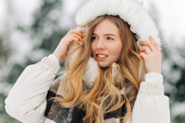 Beautiful woman standing among the snowy trees in the winter forest and enjoying the first snow. Wearing a hat, a plaid scarf and a coat