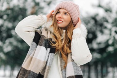 Street portrait of a beautiful happy woman in a hat posing on the street. The model looks up to the sky. Lady wearing stylish winter clothes. The magic effect of snowfall. Christmas, New Year concept