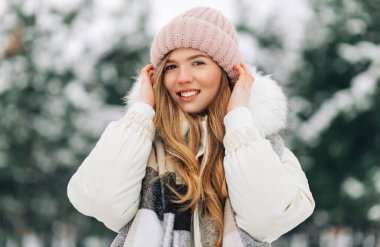 Image of a happy young woman, in a warm knitted scarf and hat looking at the camera, woman posing in the street in a snowy winter park, Winter vacation concept, winter clothes