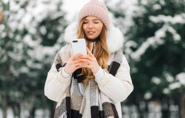 beautiful woman in a knitted hat and a warm coat and scarf walks in the winter park and uses a mobile phone. Woman in white knitted sweater. Girl using phone
