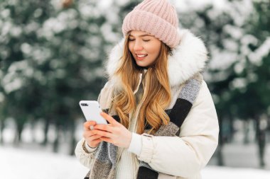 beautiful woman in a knitted hat and a warm coat and scarf walks in the winter park and uses a mobile phone. Woman in white knitted sweater. Girl using phone