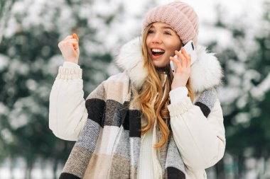 Happy excited beautiful woman in winter clothes talking on a mobile phone and celebrating victory and success, winter woman enjoying a walk in the park in winter