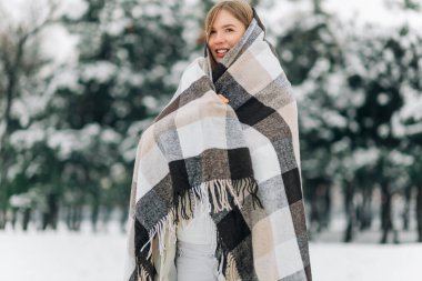 Smiling woman with warm blanket standing outdoors in winter, keeping warm. Woman covering herself with a blanket to keep warm on a snowy winter day outdoors