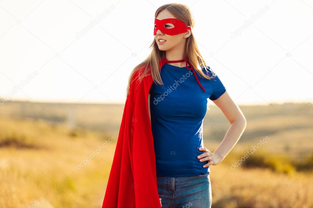 Joyful beautiful woman in a superhero costume posing against the backdrop of a sunset in nature. Superhero woman. Young and beautiful blonde in the image of a superheroine with a red cloak