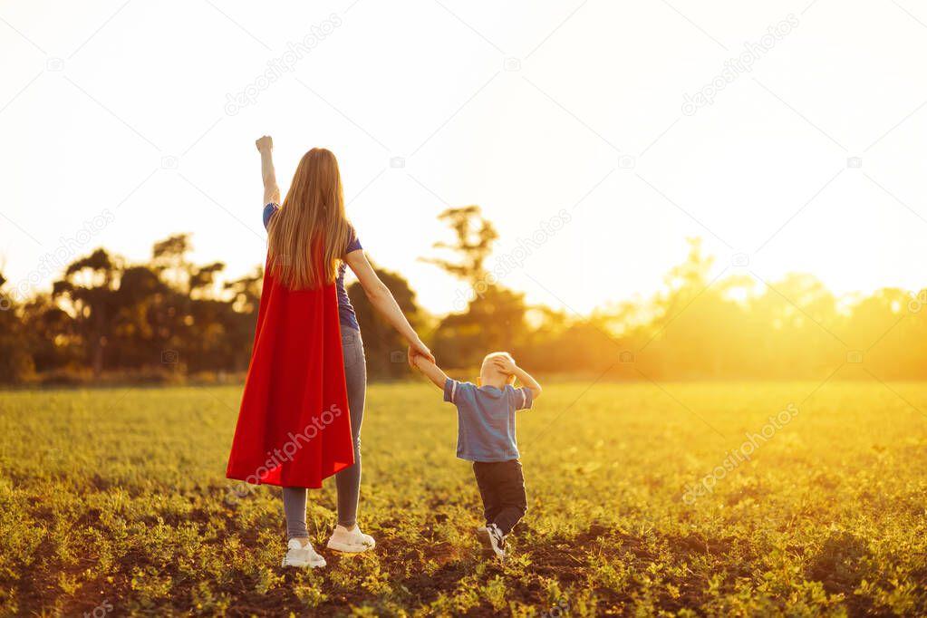 Super mom and her son walk forward holding hands. Cheerful family, a woman in a red raincoat as a superhero. Mom and son play superheroes during the day. Friendly family concept.