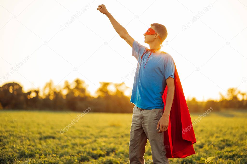 Joyful courageous man in a superhero costume poses on the field against the backdrop of sunset. A man in a red cape of a superhero makes a victory gesture, a man in the image of a superhero grows up