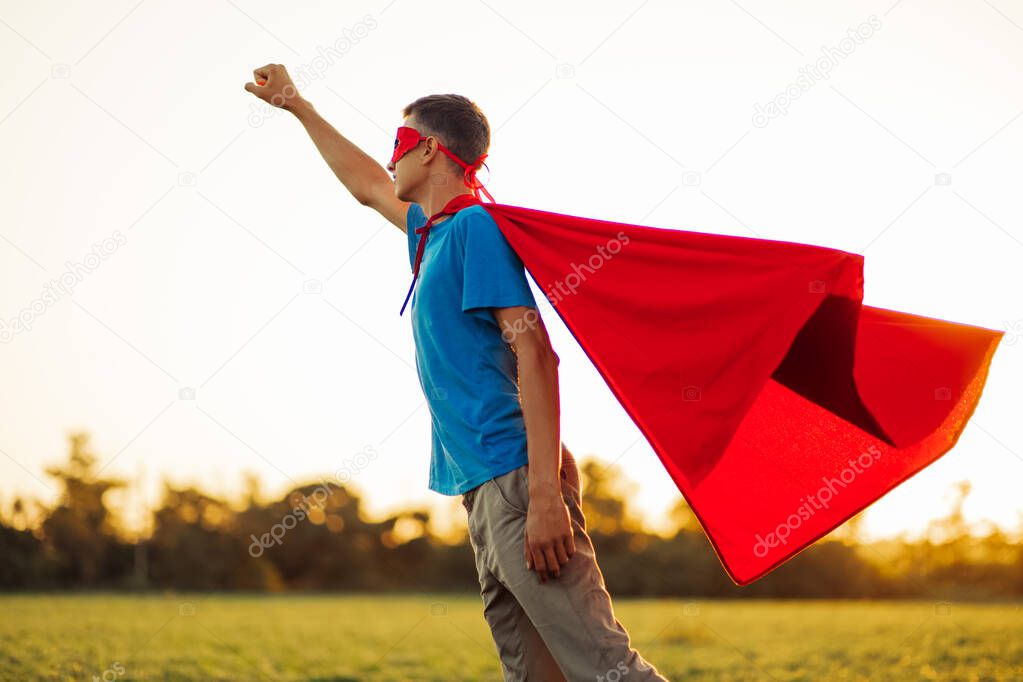 Joyful courageous man in a superhero costume poses on the field against the backdrop of sunset. A man in a red cape of a superhero makes a victory gesture, a man in the image of a superhero grows up
