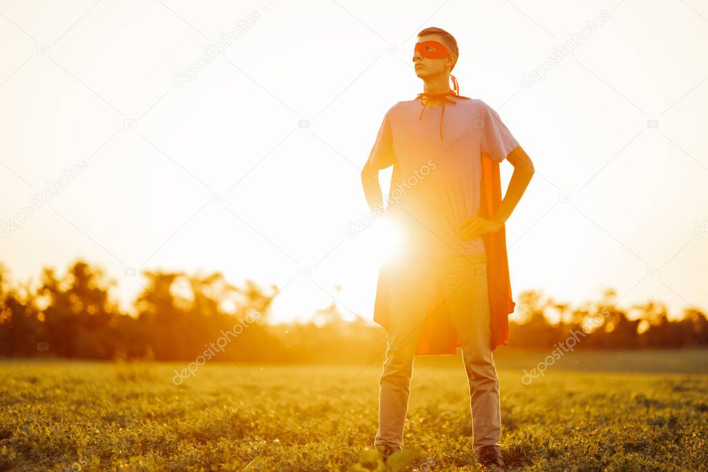 Happy man in a red cape of a superhero and a mask on his face, Strong confident man against the backdrop of a sunset sky in nature. concept of power, strength and people