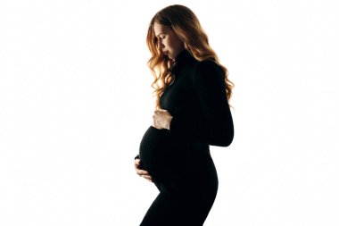 Siyah elbiseli, uzun kıvırcık saçlı güzel hamile bir kadın. Modaya uygun hamile bir anne beyaz bir arka planda karnına sarılıyor. Hamile kadınlar için moda konsepti, annelik.