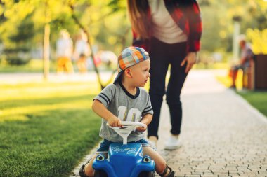 Mutlu şirin çocuk bir çocuğun arabasına biniyor, annesiyle parkta yürüyor, anne oğluna yaz günü parkta araba sürmeyi öğretiyor.