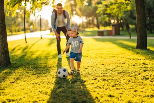 father with a little son plays football on the green grass in the park. Happy family having fun and playing football on a green grassy lawn on a sunny day. Family concept, Father\'s Day