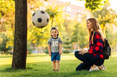 Genç mutlu anne ve küçük oğlu parkta futbol oynuyorlar, anne oğluna futbol topu veriyor, eğleniyor ve güneşli bir yaz gününde topla oynuyor.
