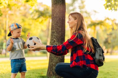 Genç mutlu anne ve küçük oğlu parkta futbol oynuyorlar, anne oğluna futbol topu veriyor, eğleniyor ve güneşli bir yaz gününde topla oynuyor.