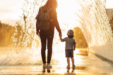Gün batımında genç bir anne ve küçük oğlunun silueti. Aile yürüyüşü, annesinin elini tutan küçük çocuk, parkta aile yürüyüşü. Aile kavramı, anneler günü.