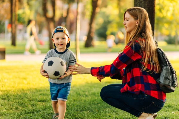 Young happy mom with little son play football in the park, mom gives her son a soccer ball, has fun and plays with the ball on a summer sunny day