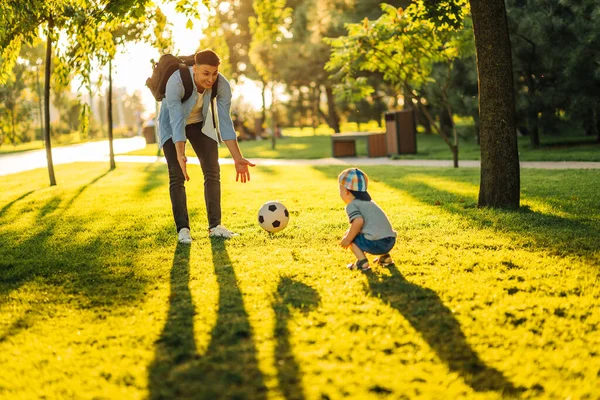 father with a little son plays football on the green grass in the park. Happy family having fun and playing football on a green grassy lawn on a sunny day. Family concept, Father\'s Day