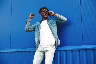 Cep telefonuyla konuşan, başarıyı ve zaferi kutlayan, evet işareti yapan, iyi haberlerden heyecanlanan kıvırcık saçlı, mavi duvarlı Amerikalı adamın yakın çekimi.