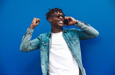 Cep telefonuyla konuşan, başarıyı ve zaferi kutlayan, evet işareti yapan, iyi haberlerden heyecanlanan kıvırcık saçlı, mavi duvarlı Amerikalı adamın yakın çekimi.