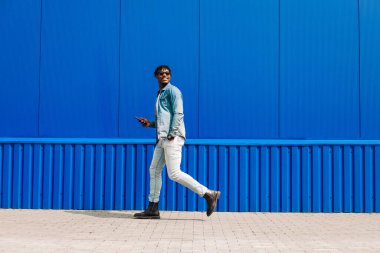 Neşeli, şık bir Afro-Amerikan erkeğinin boyu tam gelişmiş, akıllı telefon kullanıyor ve işe giderken mavi bir duvarın yanından geçiyor. Adam elinde tutuyor, cep telefonu kullanıyor, mesaj atıyor.