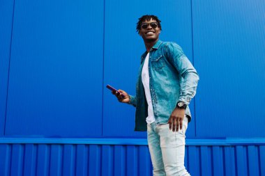 Neşeli, şık bir Afro-Amerikan erkeğinin boyu tam gelişmiş, akıllı telefon kullanıyor ve işe giderken mavi bir duvarın yanından geçiyor. Adam elinde tutuyor, cep telefonu kullanıyor, mesaj atıyor.