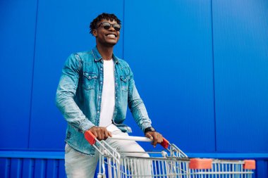 Güneş gözlüğü takan, alışveriş arabasıyla koşan ve kameraya gülümseyen şık bir Amerikalı. Yakışıklı bir adam şehirde alışveriş yapıyor ve alışveriş merkezinin yanında yürürken gülümsüyor. Alışveriş, tüketim konsepti