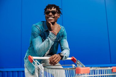 Güneş gözlüğü takan, alışveriş arabasıyla koşan ve kameraya gülümseyen şık bir Amerikalı. Yakışıklı bir adam şehirde alışveriş yapıyor ve alışveriş merkezinin yanında yürürken gülümsüyor. Alışveriş, tüketim konsepti