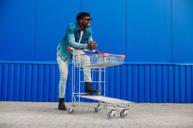 Gözlüklü, tişörtlü, kot pantolonlu ve spor ayakkabılı mutlu bir Afro-Amerikalı adamın tam vücut profili fotoğrafı. Mavi duvar arkasında alışveriş arabası olan mutlu bir adam.