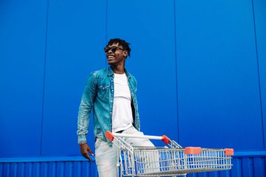 Gözlüklü, tişörtlü, kot pantolonlu ve spor ayakkabılı mutlu bir Afro-Amerikalı adamın tam vücut profili fotoğrafı. Mavi duvar arkasında alışveriş arabası olan mutlu bir adam.