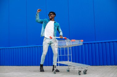 Alışveriş günü. Alışveriş arabalı mutlu siyah adamın fotoğrafı başarıyı ve zaferi kutluyor, yumruğunu kaldırıyor, açık havada mavi duvardan alışveriş yapan neşeli Afrikalı adam.