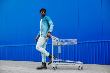 Kaygısız eğlence. Gülümseyen Afro-Amerikan alışverişçisi modaya uygun giysiler içinde, mavi bir duvarın yanında alışveriş arabasıyla. Neşeli esmer tenli hippi adam alışveriş yaptıktan sonra dinleniyor.