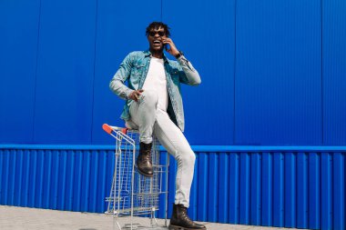 Neşeli Afro-Amerikan kıvırcık adam, güneş gözlüğü takıyor, alışveriş arabasında oturuyor ve cep telefonuyla konuşuyor, alışveriş arabasından alışveriş yapıyor, mavi arka planda. Tüketim, alışveriş