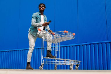 Erkek alışverişi. Alışveriş arabalı gülümseyen Afro-Amerikan adam alışverişi seviyor. Alışveriş yaparken mavi duvara karşı telefon kullanan yakışıklı bir adam. Tüketim, alışveriş, yaşam tarzı konsepti