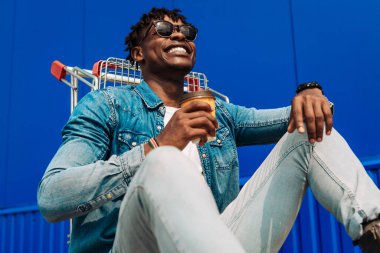 Gülümseyen Afro-Amerikalı alışverişçi alışveriş arabasında oturuyor, neşeli esmer tenli hippi adam alışveriş yaptıktan ve içtikten sonra dinleniyor, dışarıda mavi duvarda.