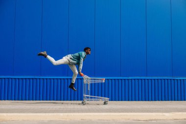 Günlük giysili ve güneş gözlüklü genç mutlu Afro-Amerikan adam dışarıda boş alışveriş arabasıyla fotokopi alanı olan mavi duvara doğru koşuyor. Alışveriş, tüketim konsepti