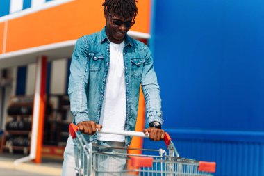 Erkek alışverişi. Gülümseyen koyu tenli, güneş gözlüklü adam alışveriş merkezinden sokakta yürüyen bir alışveriş arabasıyla çıkıyor. Tüketim, yaşam tarzı konsepti