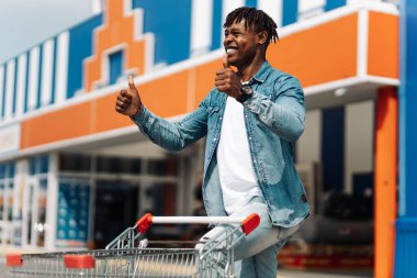 Erkek alışverişi. Güneş gözlüklü mutlu Afrikalı bir adam alışveriş merkezinden alışveriş arabasıyla çıkıyor başparmak hareketi yapıyor, alışverişi seven bir adam. Tüketim, yaşam tarzı konsepti