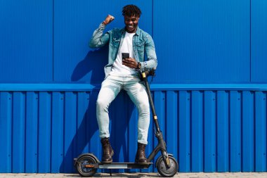 Modern binanın yakınında elektrikli scooterlı, neşeli, esmer tenli bir adam cep telefonu kullanıyor ve akıllı telefona bakıyor. Zafer jestini kutluyor.