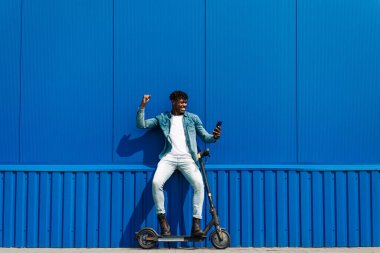 Şehirli bir sokakta elektrikli scooter kullanan heyecanlı bir Afrikalı Amerikalı. Telefonu tutan, gülümseyen, iyi haberler okurken sevinen, kazanan jestler yapan bir adam. Modern ve ekolojik ulaşım kavramı