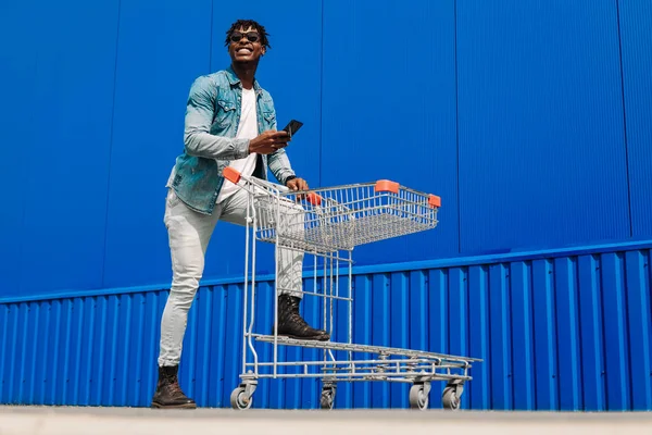 Erkek alışverişi. Alışveriş arabalı gülümseyen Afro-Amerikan adam alışverişi seviyor. Alışveriş yaparken mavi duvara karşı telefon kullanan yakışıklı bir adam. Tüketim, alışveriş, yaşam tarzı konsepti