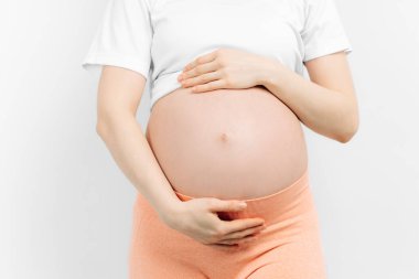 Hamile kadın ellerini beyaz bir arka planda karnının üstünde tutuyor. Hamilelik, annelik, hazırlık ve beklenti konsepti. Yakın plan, hamileliğin güzel, yumuşak ruh hali fotoğrafı..