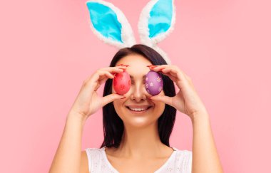 Tavşan kulaklı güzel ve pozitif esmer kadın ve boyalı Paskalya yumurtalarıyla Paskalya yumurtaları, göz, paskalya, stüdyoda pembe arka planda. Yüksek kalite fotoğraf