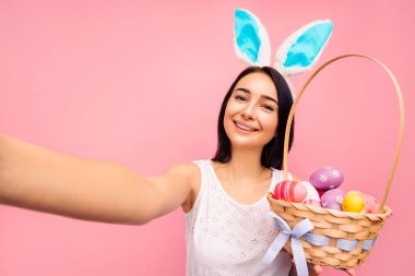 Tavşan kulaklı güzel, neşeli bir kadın selfie çekiyor, elinde yayyuami olan bir sepet tutuyor, video çağrısı, paskalya, stüdyoda pembe arka planda.