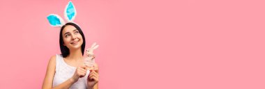 Tavşan kulaklı tatlı bir kadın elinde oyuncak bir tavşan tutuyor, geleneksel bir bayram, stüdyoda pembe bir arka planda, bir paskalya hediyesi. Yüksek kalite fotoğraf