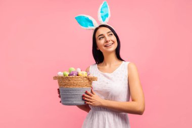 Tavşan kulaklı mutlu kadın renkli yumurtalarla dolu bir sepet tutuyor, geleneksel bir bayram, stüdyoda, pembe arka planda, Paskalya havasında. Yüksek kalite fotoğraf