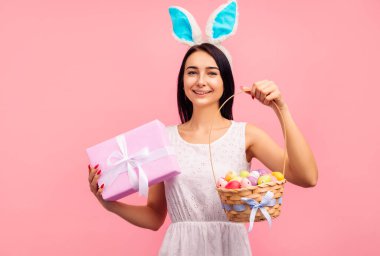 Tavşan kulaklı güzel bir kadın elinde bir sepet renkli yumurta, geleneksel bir tatil, stüdyoda pembe arka planda, Paskalya havasında.