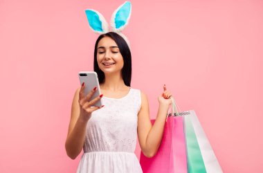 Tavşan kulaklı mutlu kadın ellerinde alışveriş torbalarıyla telefonda mesaj yazıyor, paskalya havasında.