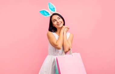 Elinde alışveriş poşetleri, bahar alışverişi, geleneksel tatil, pembe arka planda stüdyoda paskalya havasında tavşan kulaklı bir kadın.