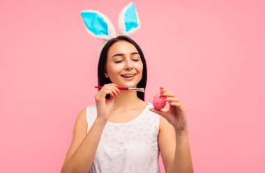 Tavşan kulaklı mutlu kadın Paskalya yumurtalarını boyuyor, Paskalya bayramını hazırlıyor, stüdyoda pembe bir arka planda, Hıristiyanlık