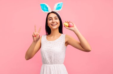 Tavşan kulaklı ve boyalı paskalya yumurtalı mutlu esmer kız pembe arka plandaki stüdyoda paskalya zaferini işaret ediyor. Yüksek kalite fotoğraf