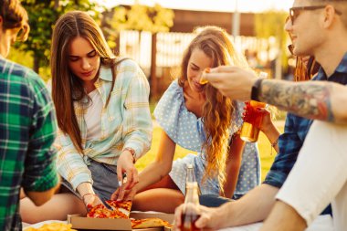 Bir grup arkadaş parkta pizzanın tadını çıkarıyorlar. Erkekler ve kadınlar pizza kutusunun etrafındaki çimlerde otururlar. Parkta piknik yapmak, doğada arkadaş olmak.